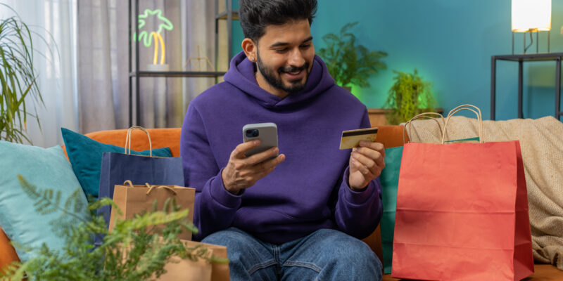 Hombre joven realizando una compra online desde su celular, mientras sostiene una tarjeta de crédito y está rodeado de bolsas de compras, representando el hábito moderno de consumir por internet desde casa.